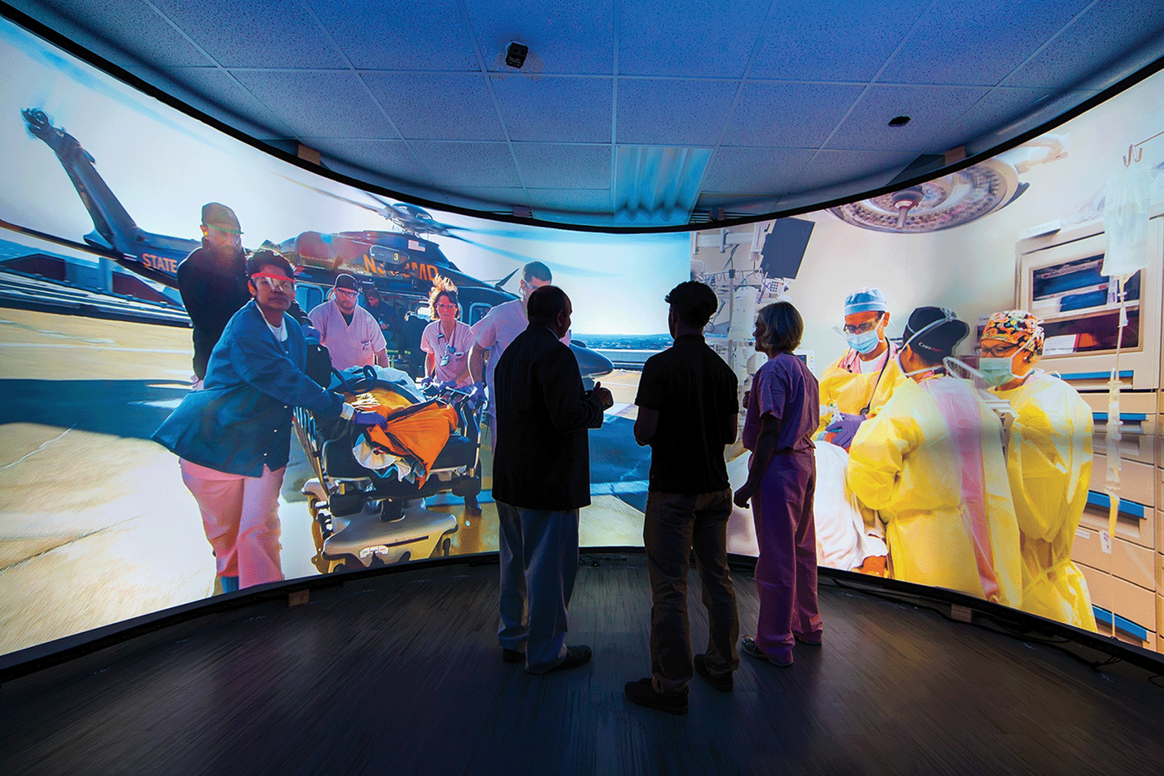 Two nursing students and a teacher view an immersive curved screen displaying vivid scenes of emergency medical care and surgery
