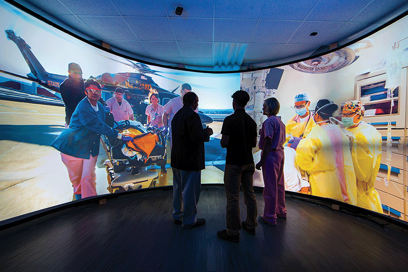 Two nursing students and a teacher view an immersive curved screen displaying vivid scenes of emergency medical care and surgery