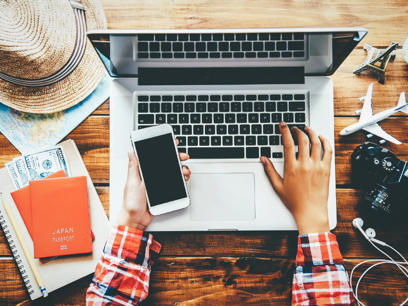 A man sits at a laptop with a cell phone in his hand and travel items on the table around him