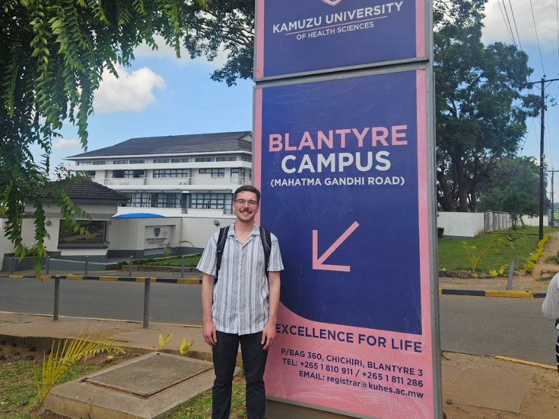 A student stands outside beside a sign for Kamuzu's Blantyre Campus