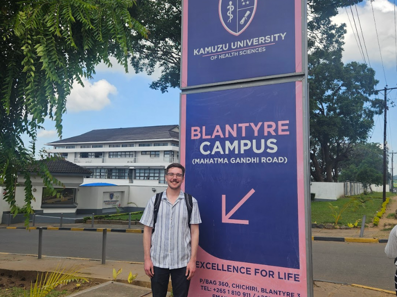 A student stands outside beside a sign for Kamuzu's Blantyre Campus
