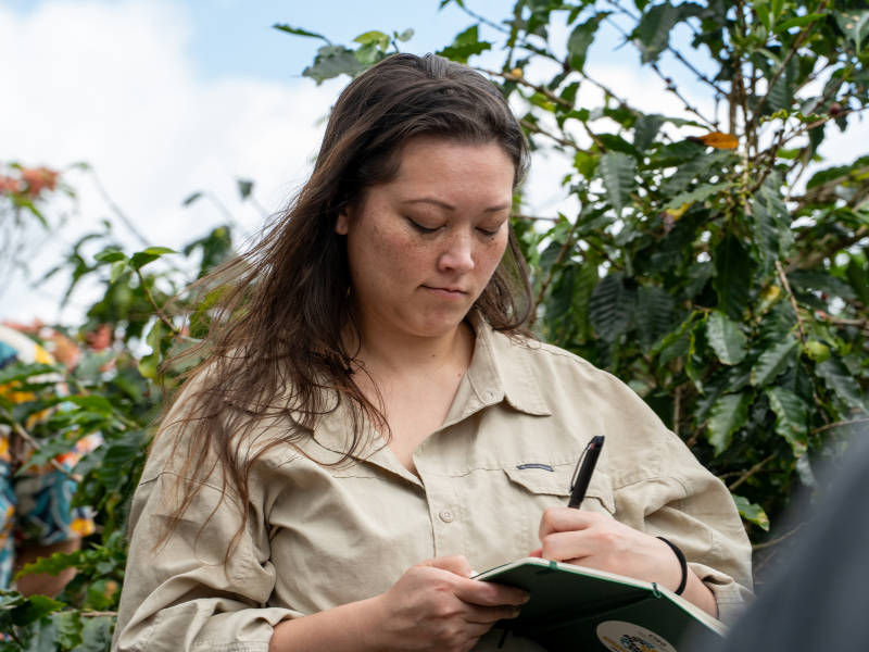 A woman writes in a journal outside with bushes behind her