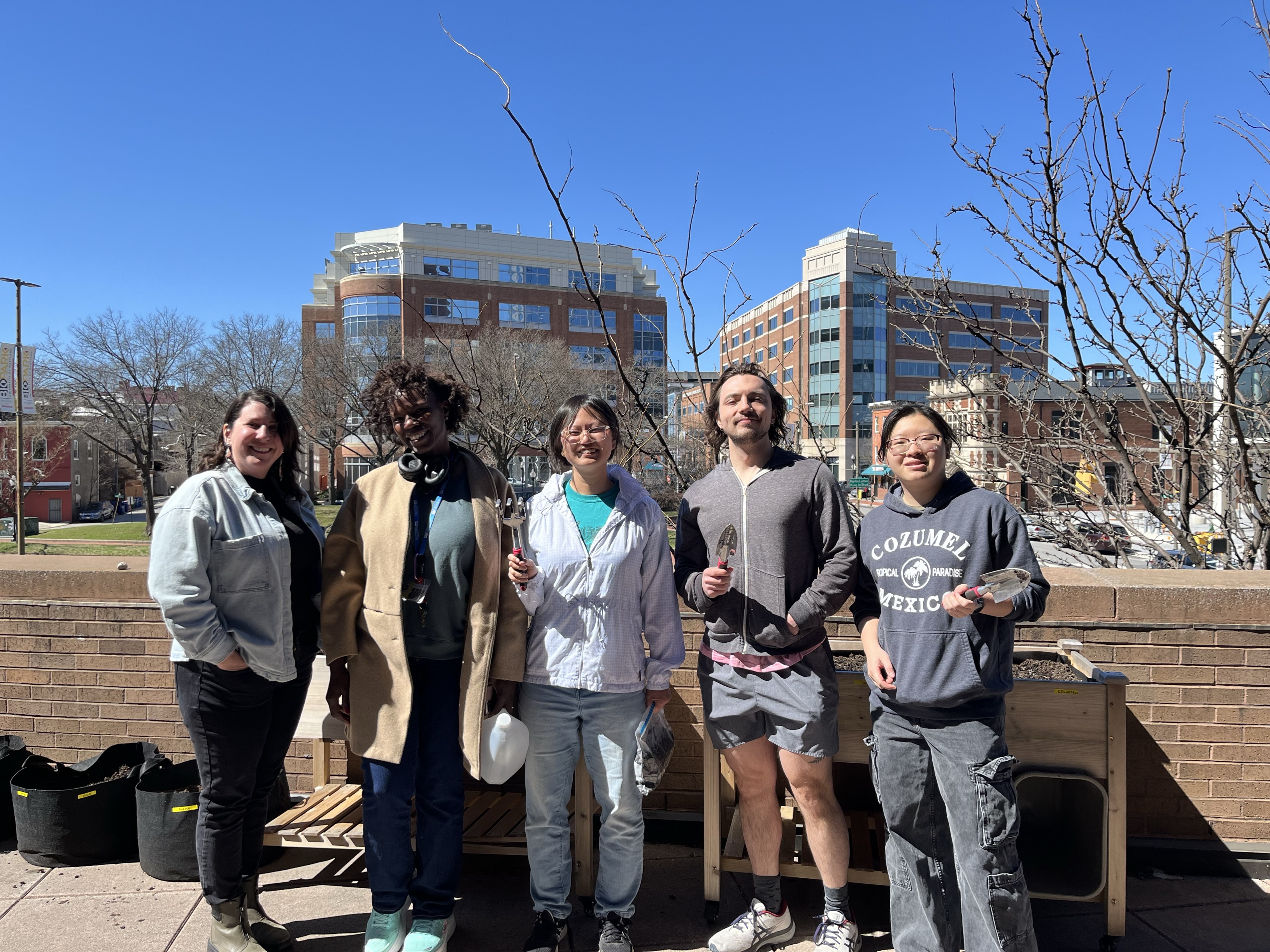 5 people standing in a row, outside in front of the community garden. 